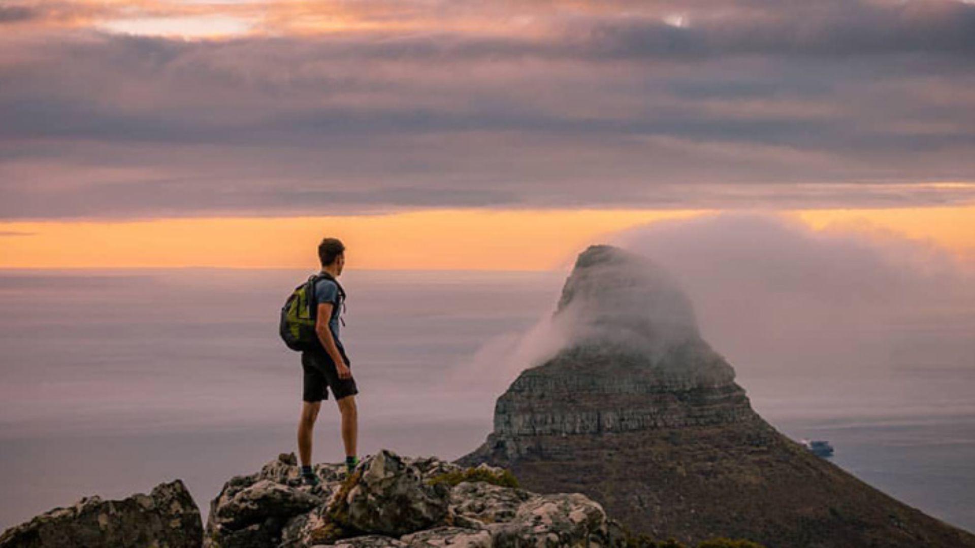 A man stands atop a mountain, wearing a backpack, gazing at the expansive landscape below.