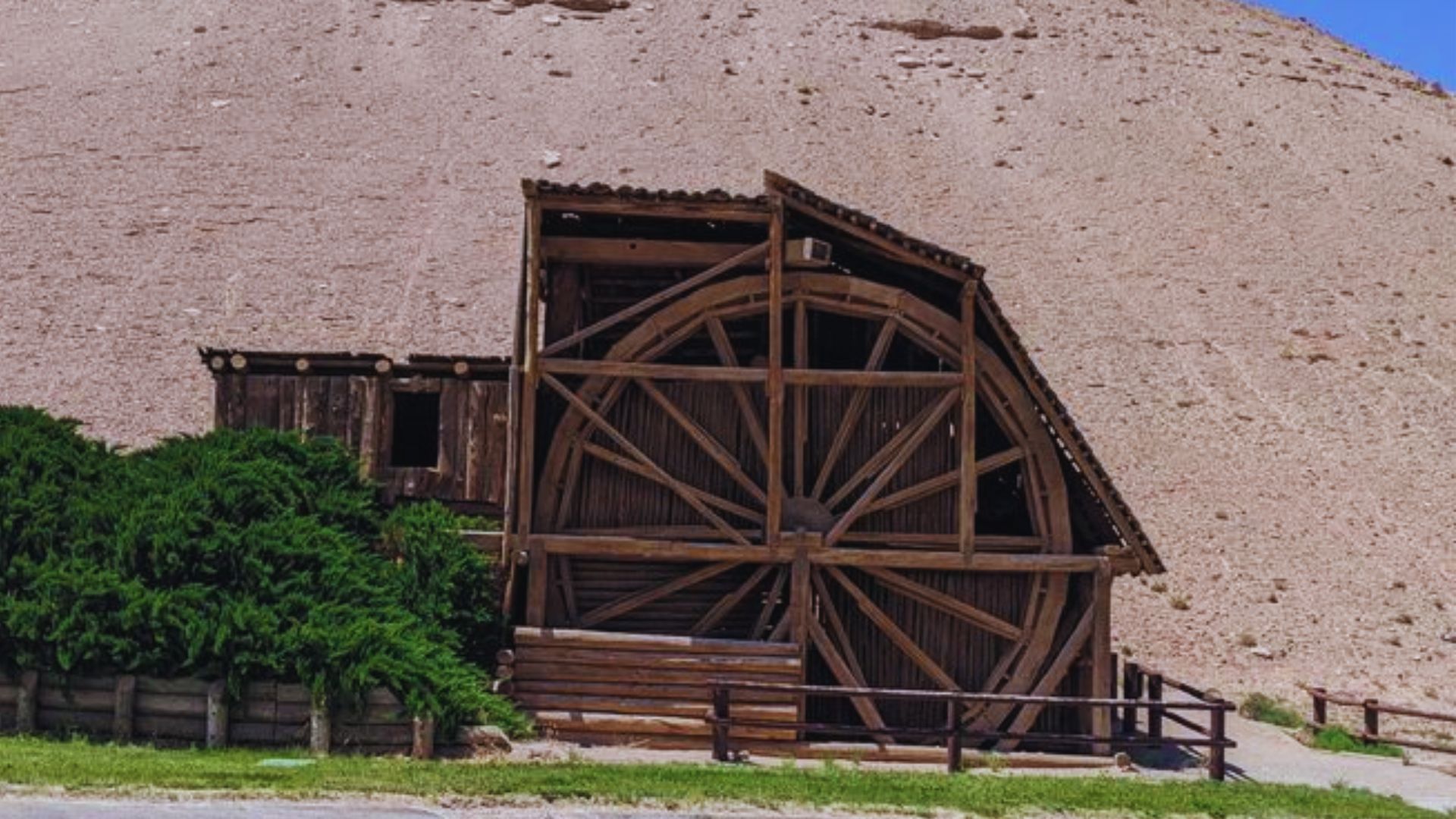 A large wooden water wheel stands in front of a steep hill, showcasing a blend of nature and traditional craftsmanship.