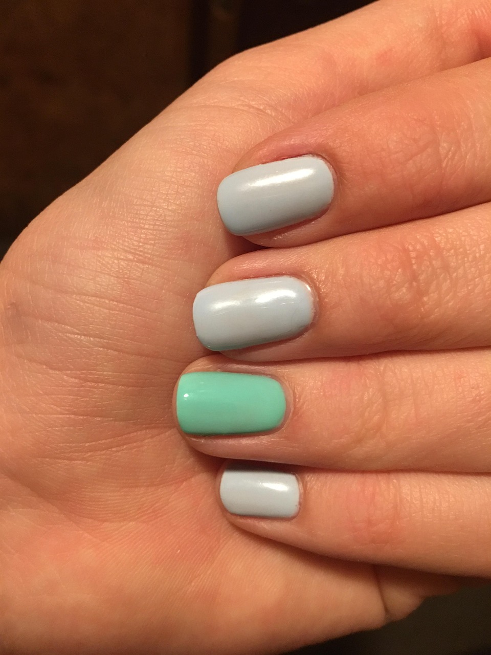 A hand with manicured nails, four painted light blue and one in mint green. The lighting is warm, highlighting a soft, relaxed ambiance.