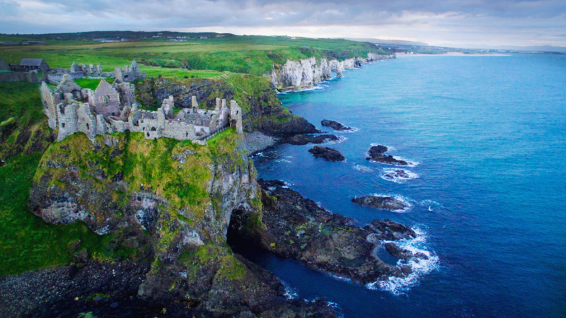 Aerial view of Dunluce Castle ruins on a lush green cliffside, overlooking the deep blue ocean. Rugged coastline stretches under a cloudy sky.
