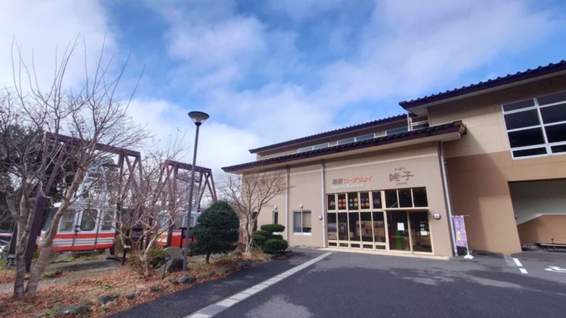 A modern beige building with large windows is beside a garden with bare trees and shrubs. A vintage train car is visible on the left. The sky is partly cloudy.