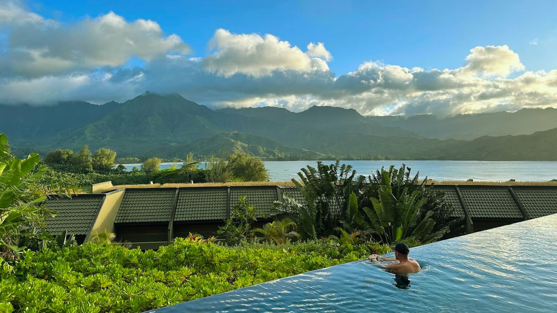 A person relaxes in an infinity pool, overlooking lush greenery and distant mountains under a partly cloudy sky, creating a serene and tranquil scene.