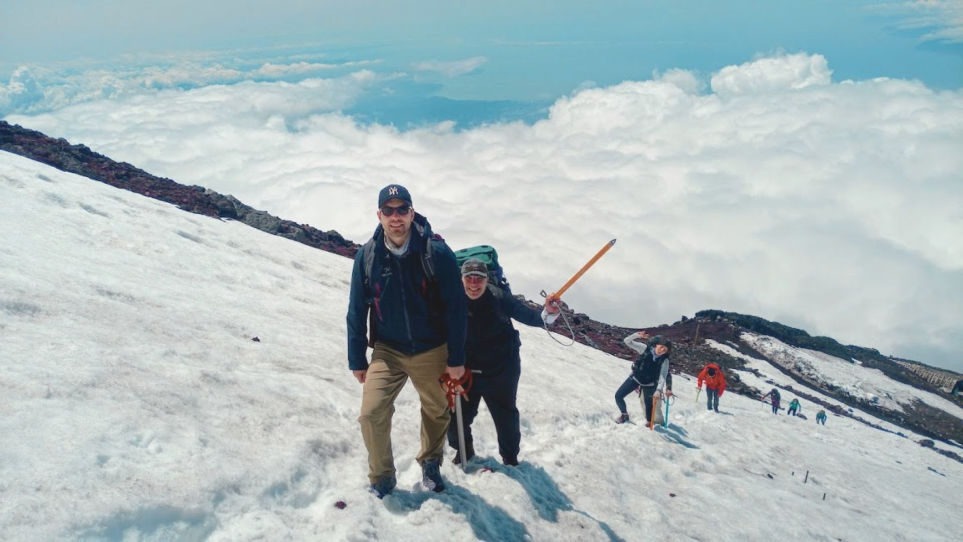 A group of climbers ascends a snowy mountainside beneath a bright sky, smiling and using hiking poles, with expansive clouds below them, conveying adventure.