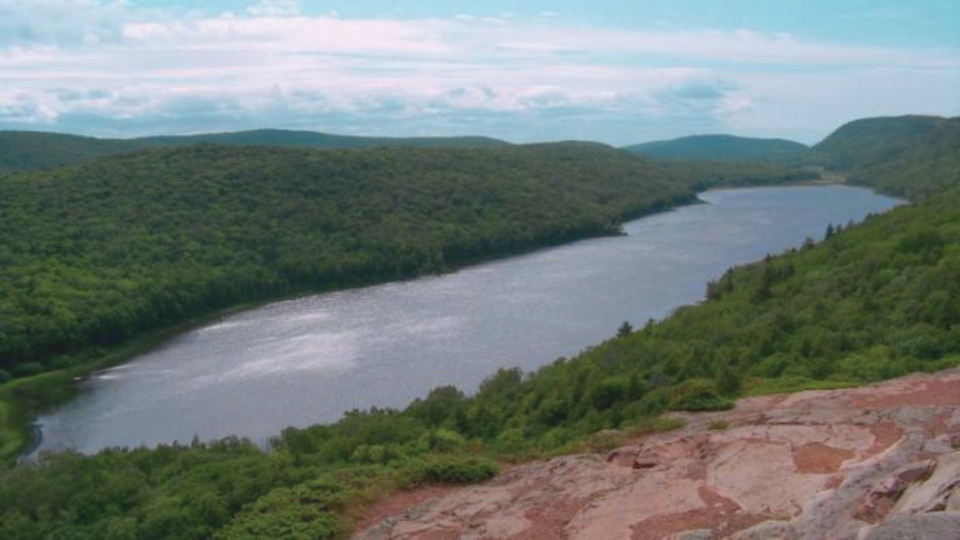 Expansive view of a serene lake snaking through lush, green hills under a bright blue sky with scattered clouds, evoking a sense of tranquility and natural beauty.