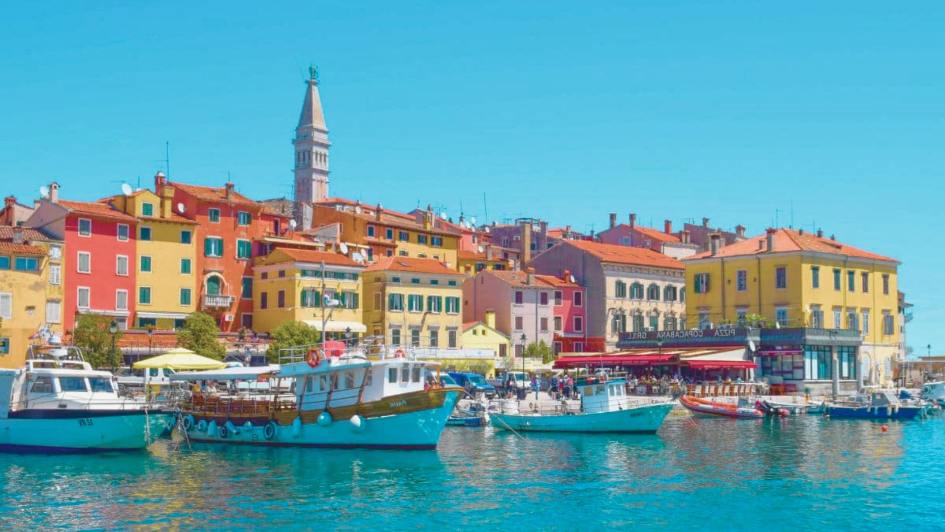 A vibrant coastal town scene with colorful buildings and boats on clear blue water under a bright sky. The atmosphere is lively and inviting.
