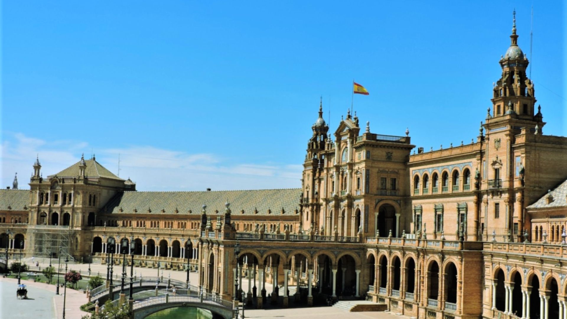 Grand plaza with ornate, arched brick buildings under a clear blue sky. Spanish flag atop central tower. People stroll, enhancing lively atmosphere.
