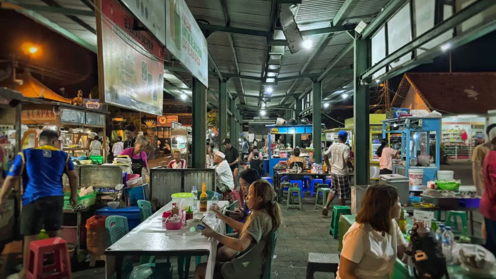A bustling night market with people dining at tables under a covered area. Food stalls line each side, creating a lively, social atmosphere.