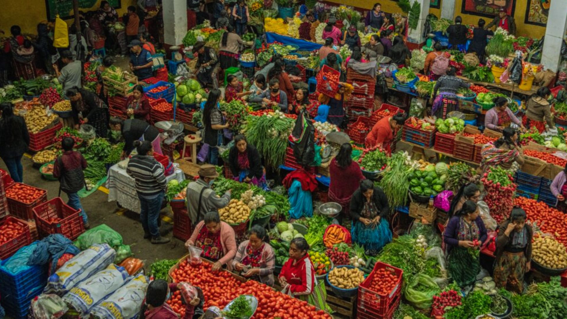 Bustling indoor market with vibrant piles of fruits and vegetables. People in colorful clothing engage in lively trade, creating a lively, dynamic scene.