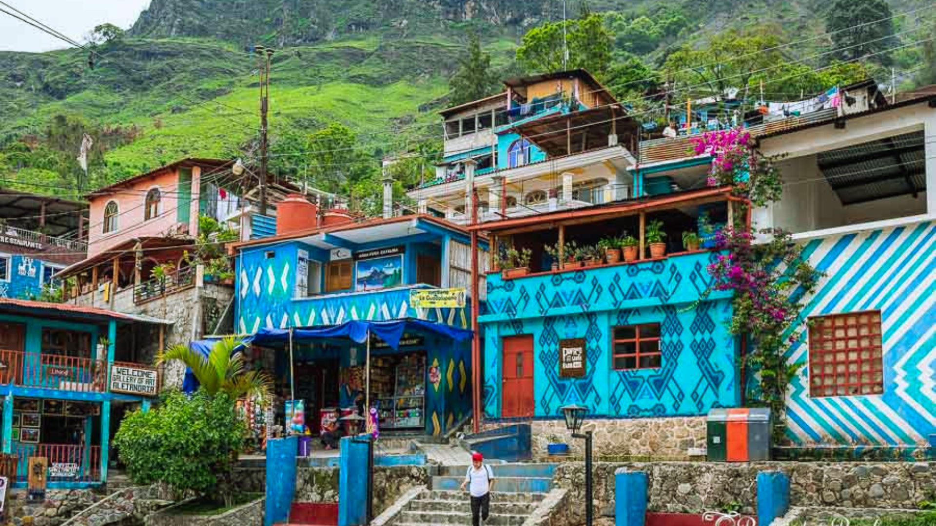 Colorful hillside houses in vibrant blues and reds, adorned with geometric patterns and lush greenery. A person walks down stone steps in the foreground.