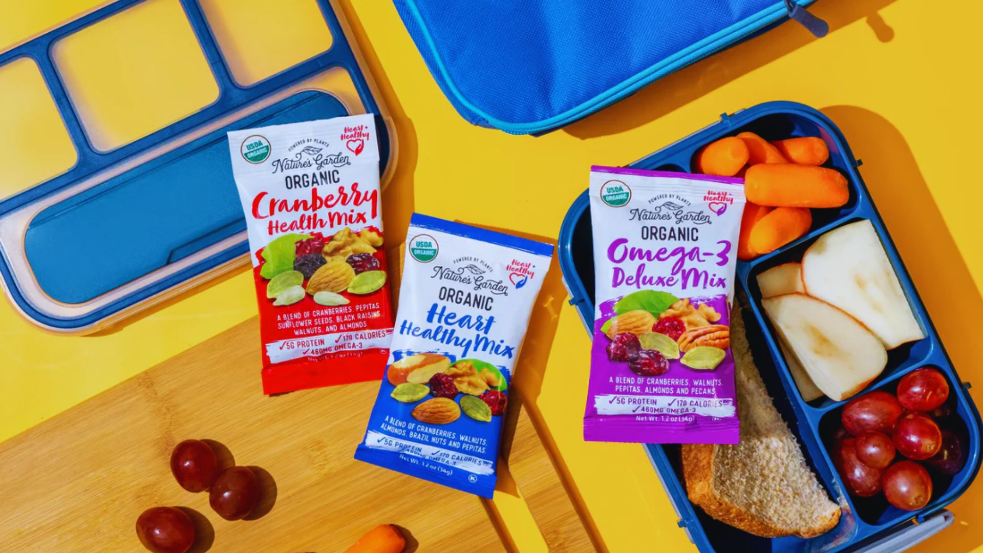 A vibrant lunch setup features healthy snacks with three colorful Nature's Garden organic mix packets on a yellow background. A lunchbox contains carrots, apple slices, grapes, and a sandwich, conveying a wholesome, nutritious vibe.