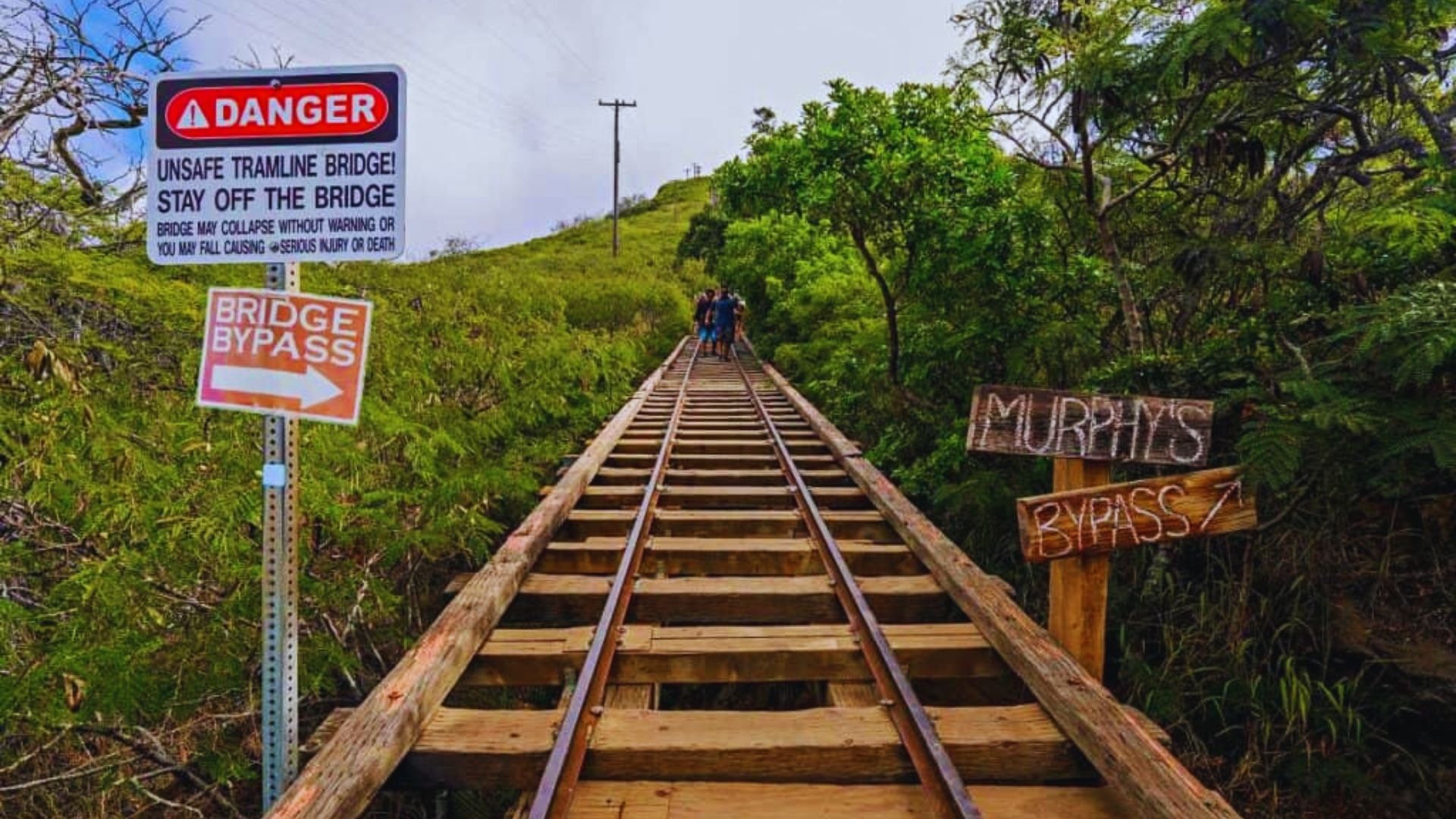 An old, unsafe tramline bridge set in lush greenery with warning signs. People in the distance walk cautiously, highlighting a sense of adventure.