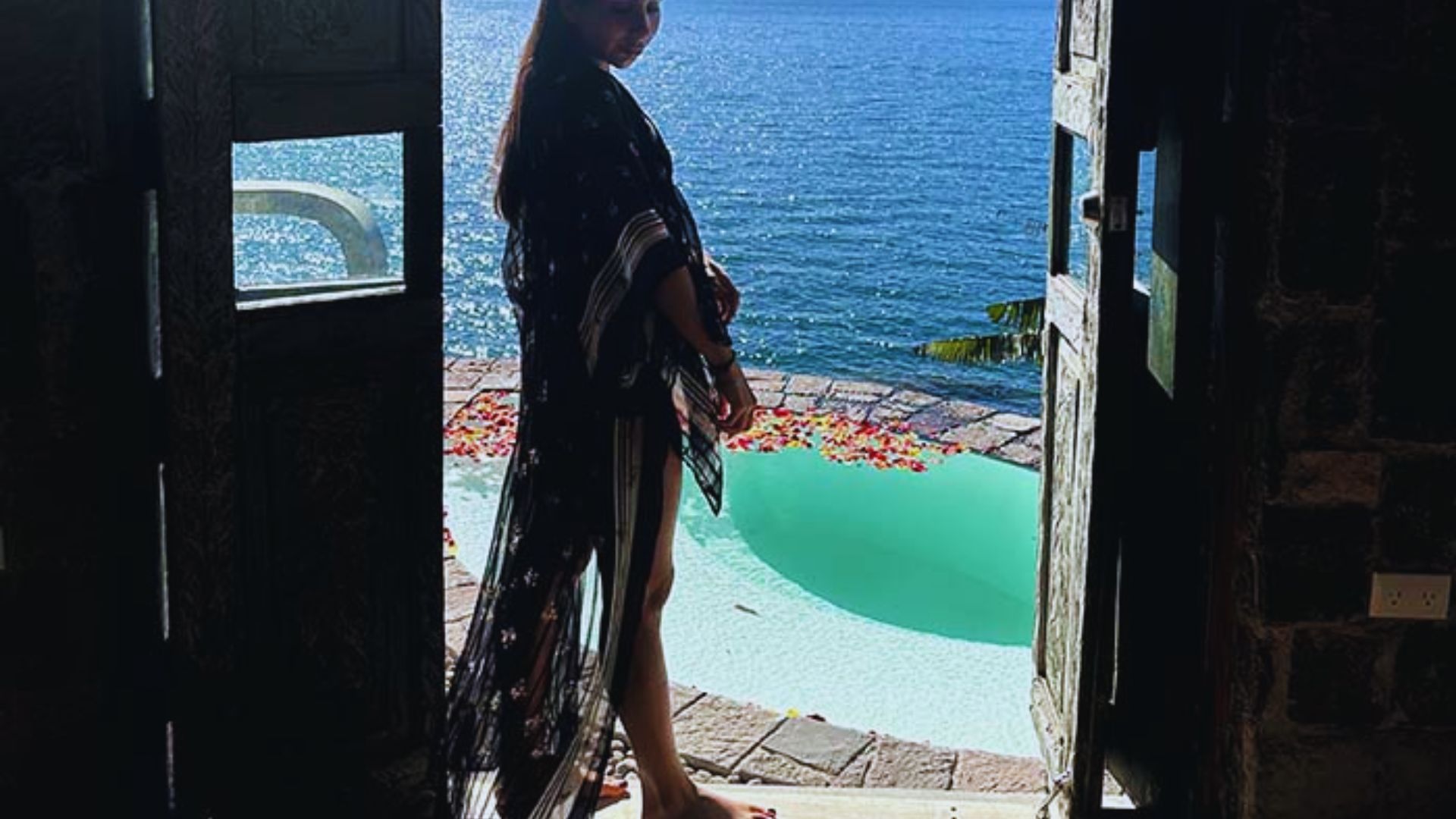 A woman in a black dress stands elegantly by a pool, reflecting the serene atmosphere of the setting.