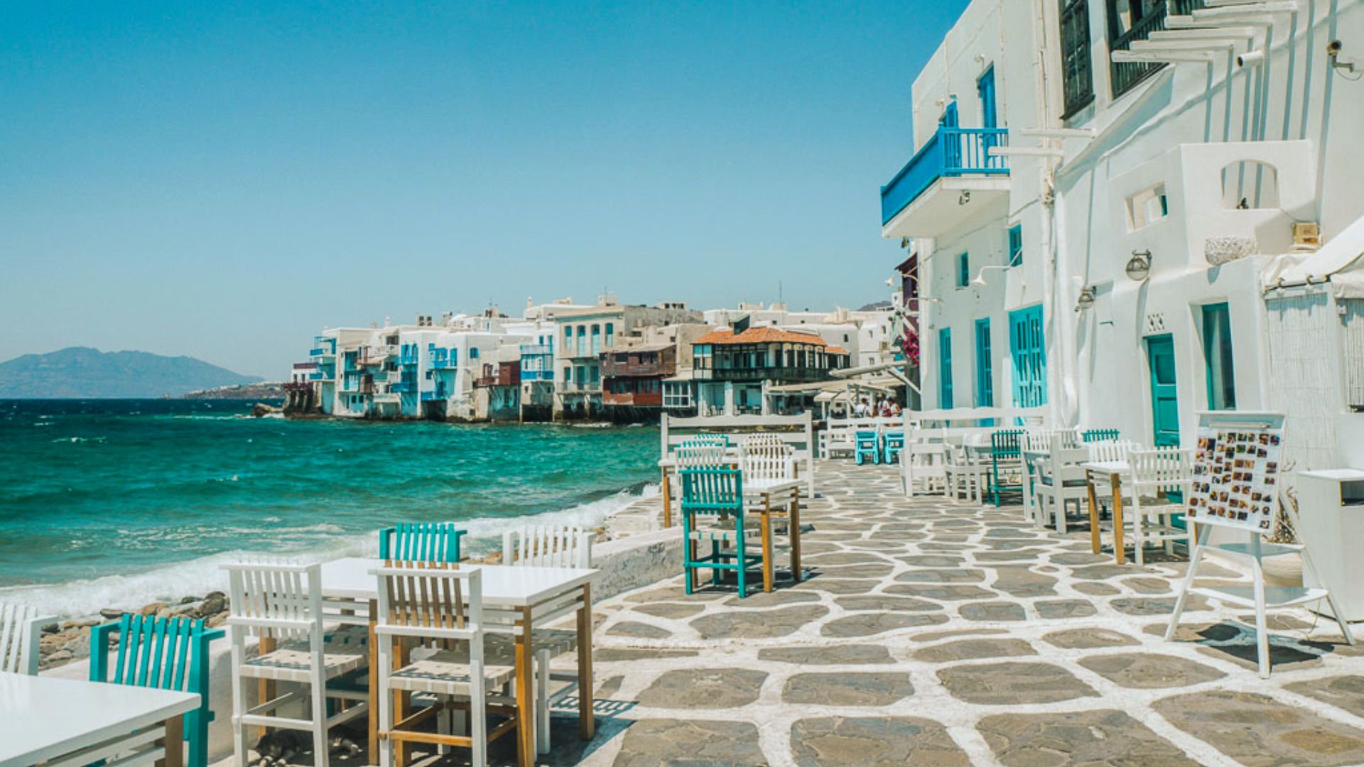 Seaside scene in Mykonos with white and blue buildings, turquoise sea, and an empty waterfront cafe with white tables and chairs on a sunny day.