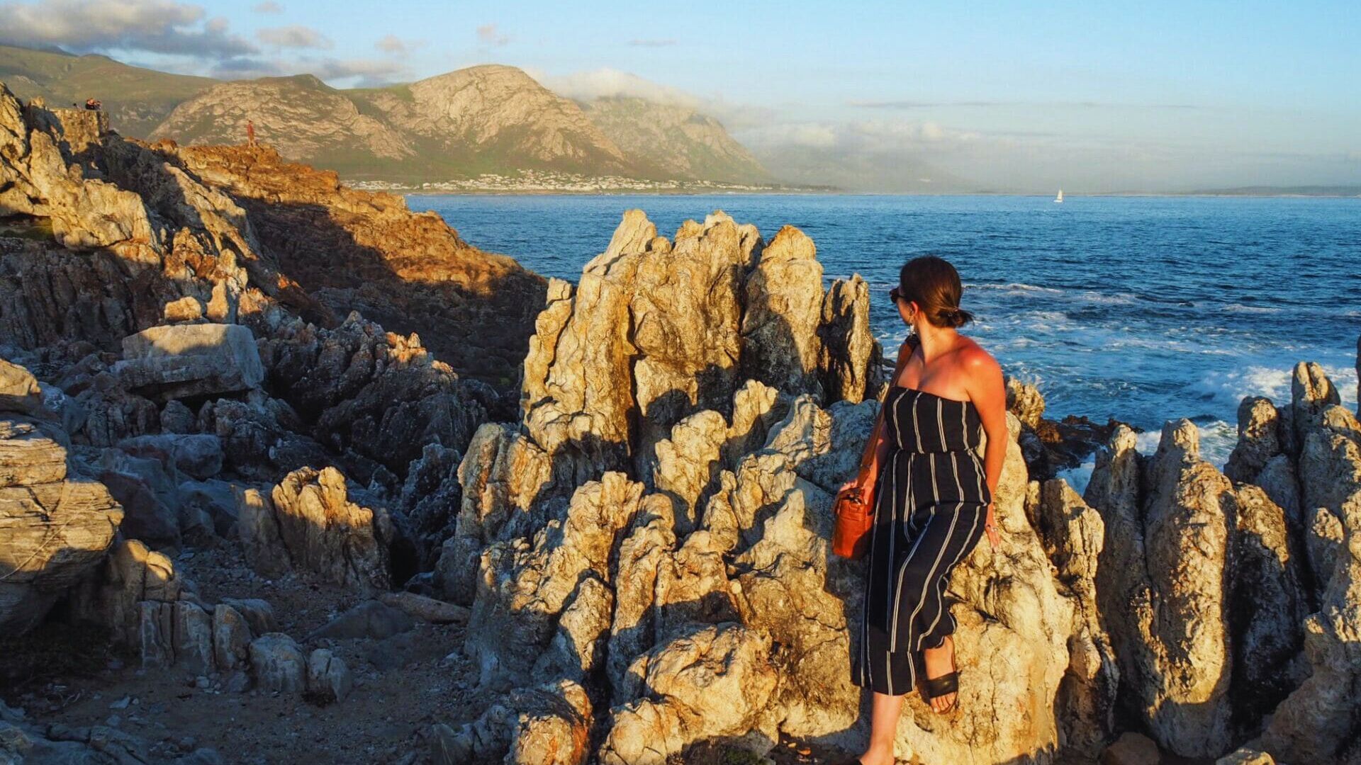 A woman stands on rocky terrain, gazing out at the vast ocean under a clear blue sky.