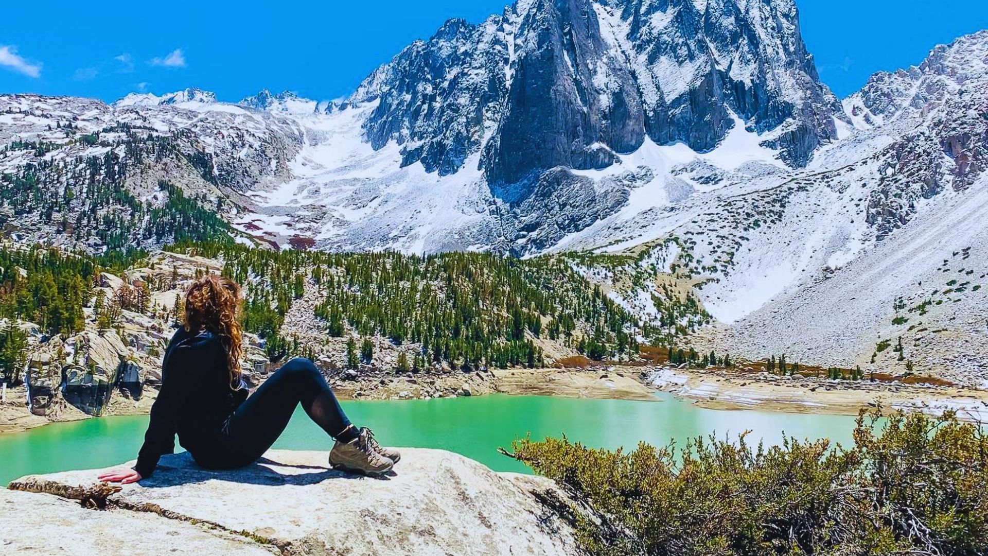 A woman relaxes on a rock, admiring the calm lake and scenic landscape under bright sunlight.