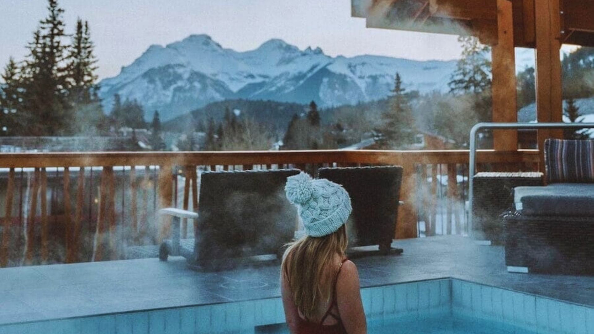 A woman wearing a hat relaxes in a hot tub, with majestic mountains visible in the background.