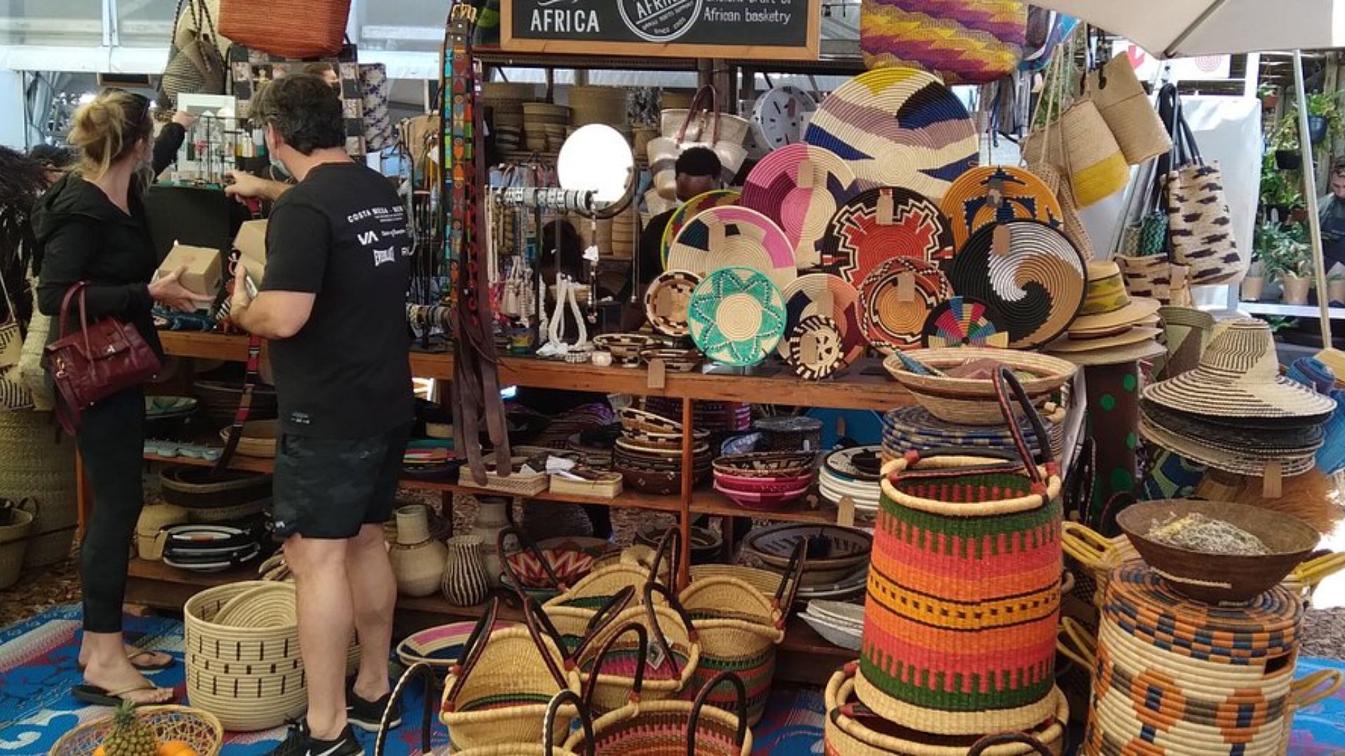 A bustling market scene with shoppers carrying baskets and various items, surrounded by colorful stalls and products.