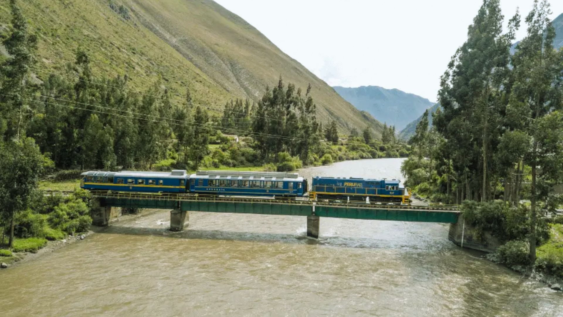 A blue train crosses a narrow bridge over a swift river, surrounded by lush green mountains and tall trees under a cloudy sky.