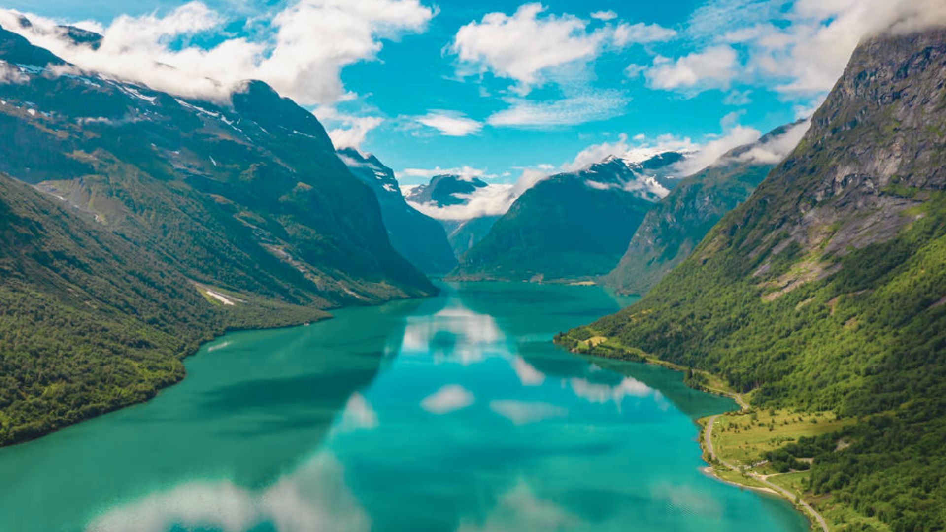 A serene fjord view with clear turquoise water reflecting the sky. Snow-capped mountains and lush green hills frame the scene under a bright blue sky.