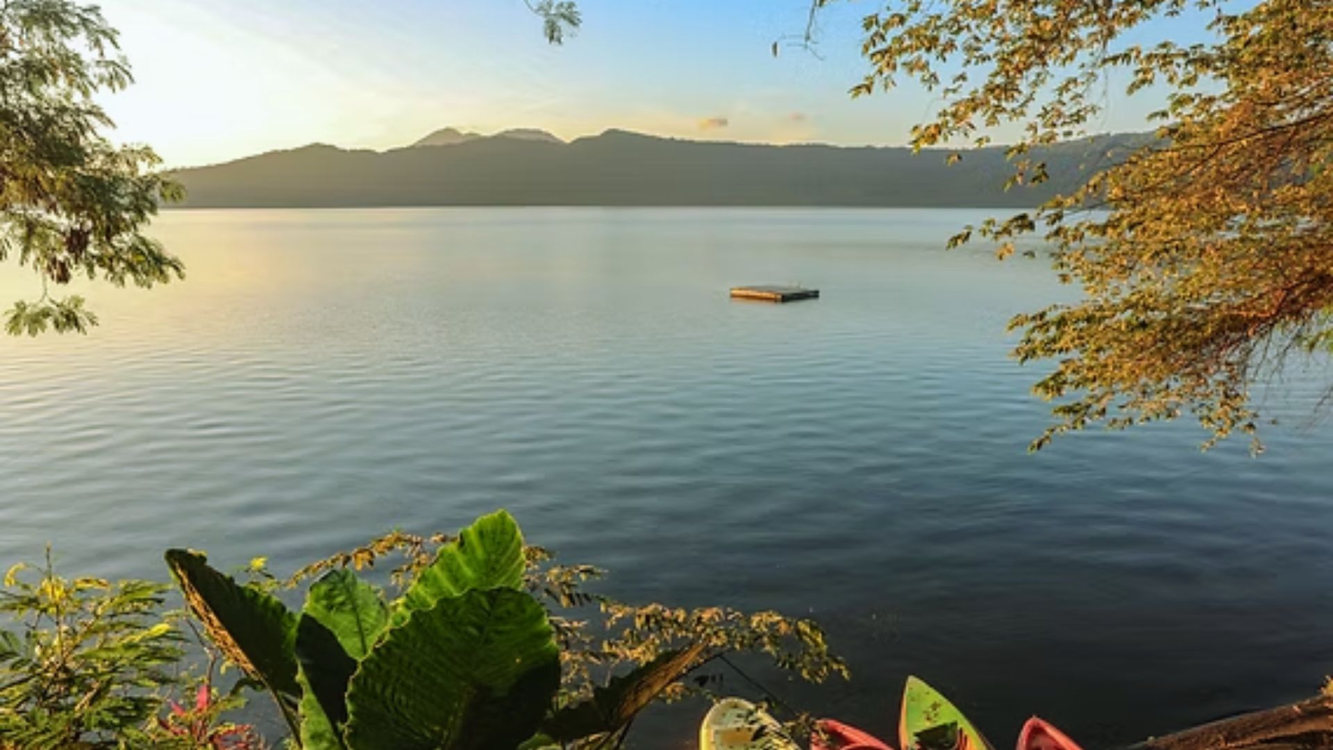 Calm lake at sunrise with a small floating dock, framed by lush green foliage and trees. Distant mountains under a clear sky create a serene atmosphere.