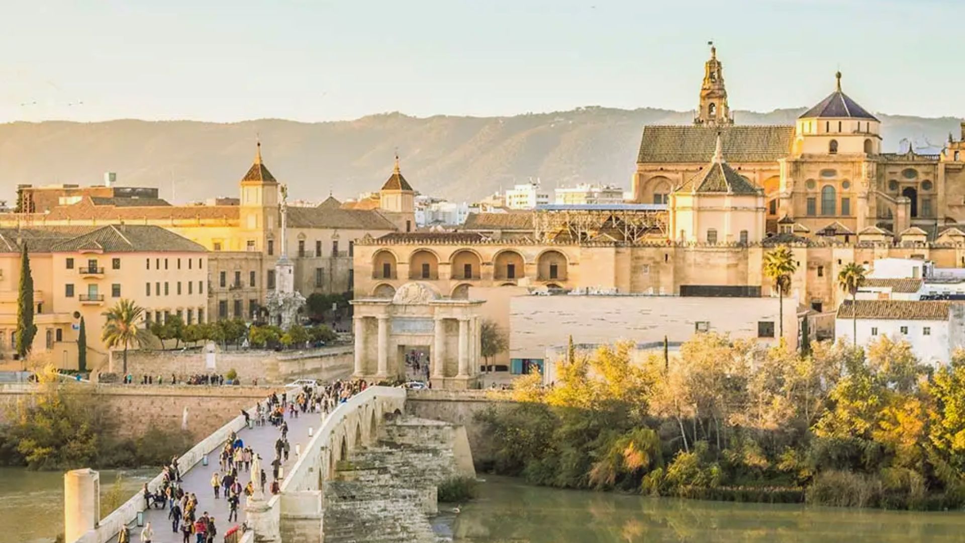 A sunlit view of Córdoba, Spain, featuring the ancient Roman bridge and the historic Mosque-Cathedral. Mountains in the background enhance the scene's serenity.