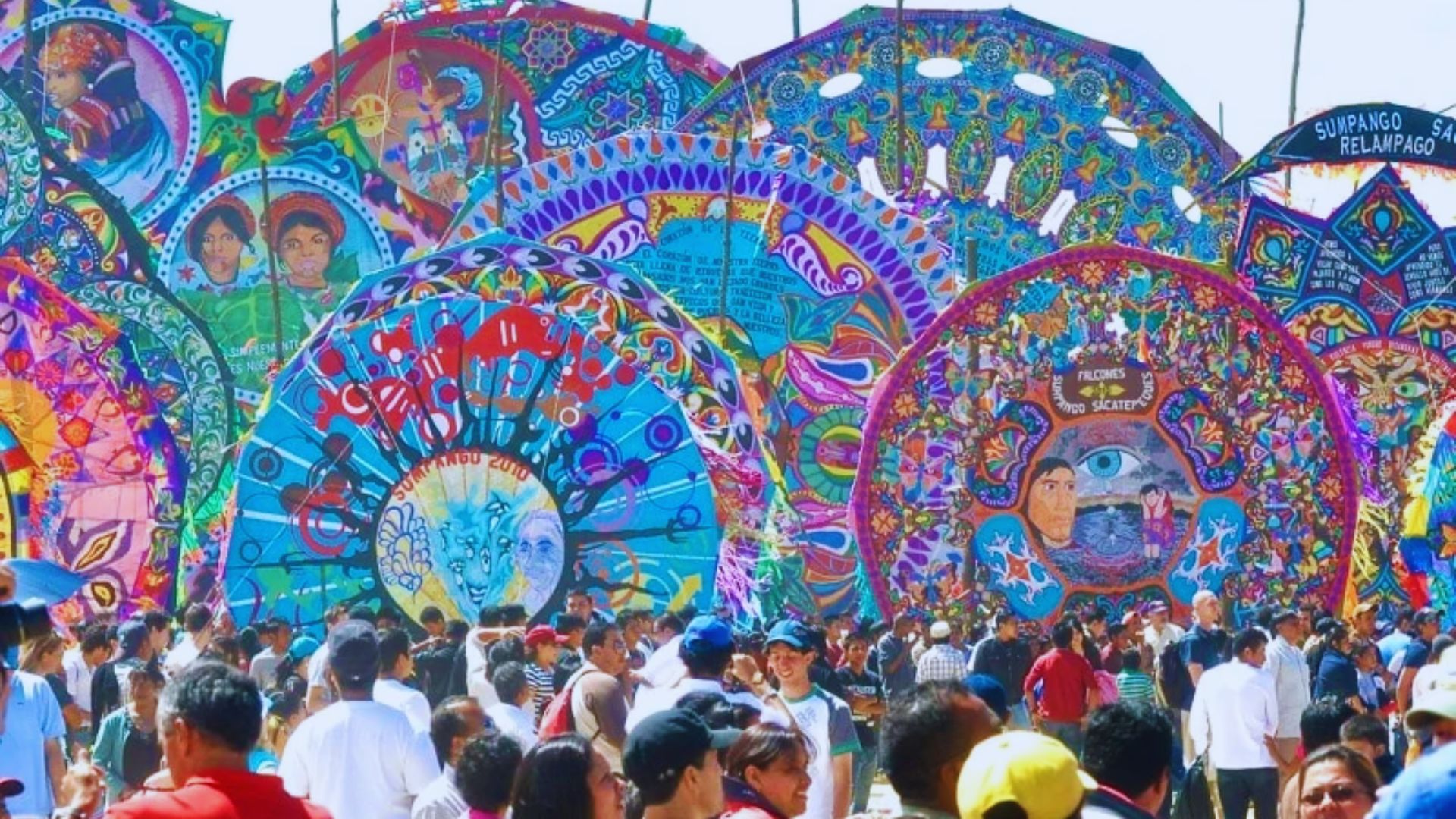 A vibrant festival scene with a crowd of people in front of large, colorful kites adorned with intricate patterns and faces, creating a lively atmosphere.