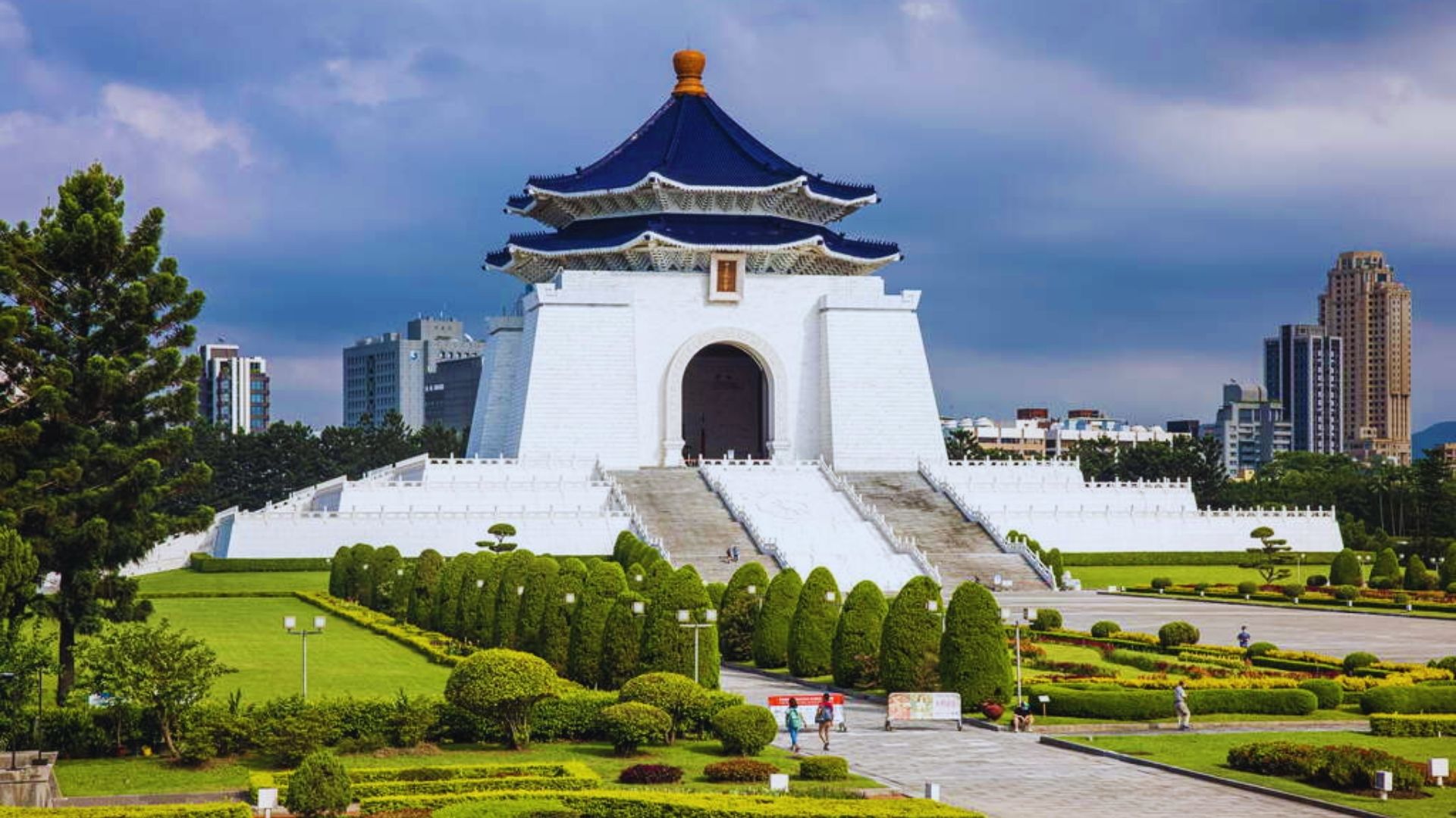 A majestic white memorial hall with a blue-tiled pagoda roof stands amid lush gardens and manicured hedges under a cloudy sky, with city skyscrapers in the distance.