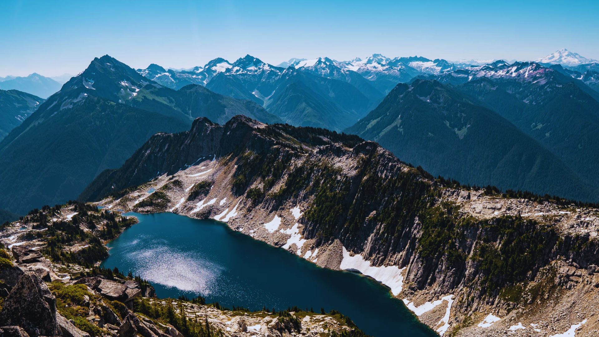 A serene mountain landscape featuring a clear blue alpine lake surrounded by rocky cliffs, with snow-capped peaks in the distance under a bright blue sky.