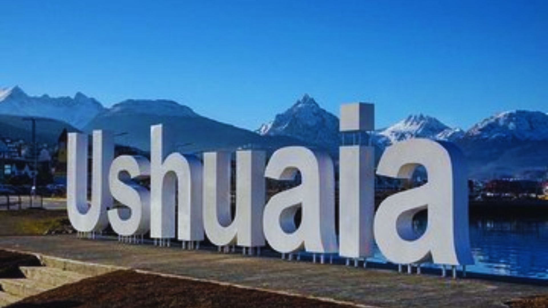 Large white letters spelling "Ushuaia" stand against a backdrop of snow-capped mountains and a clear blue sky, conveying a sense of serene, natural beauty.