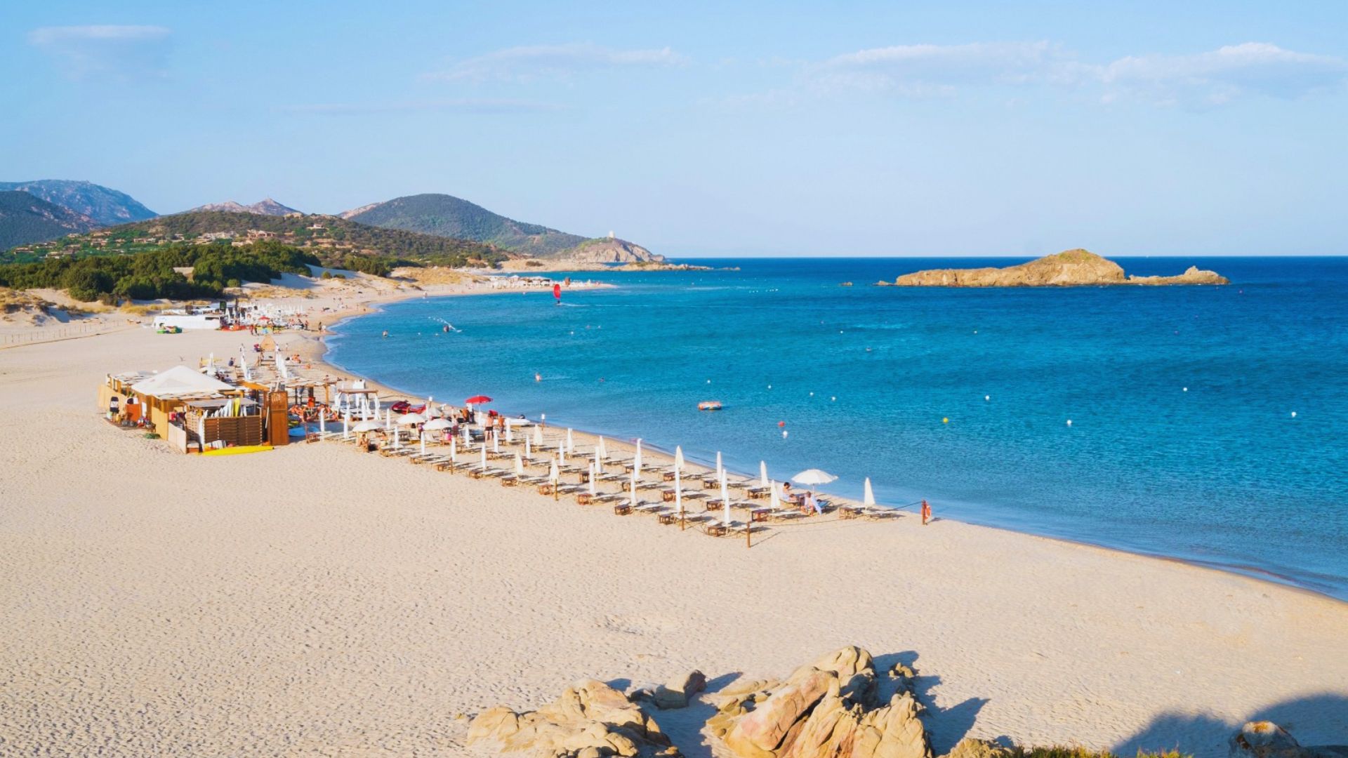 Sunny beach scene with clear blue water and gentle waves. Rows of white umbrellas line the sandy shore, with distant green hills and rocks.