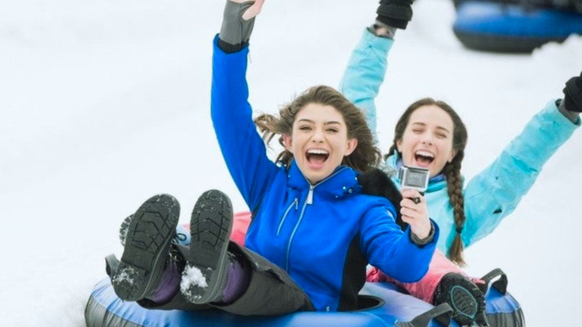 Two people joyfully snow tubing down a slope. Both wear bright winter jackets, arms raised, with wide smiles, capturing a carefree, exhilarating moment.