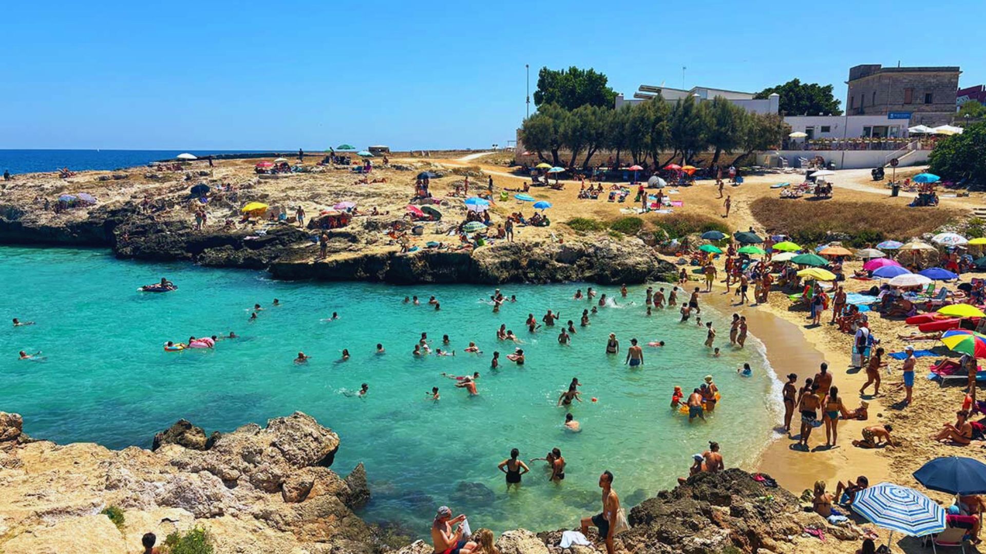 A busy beach scene with numerous people swimming in the water and sunbathing on the sand under bright sunlight.
