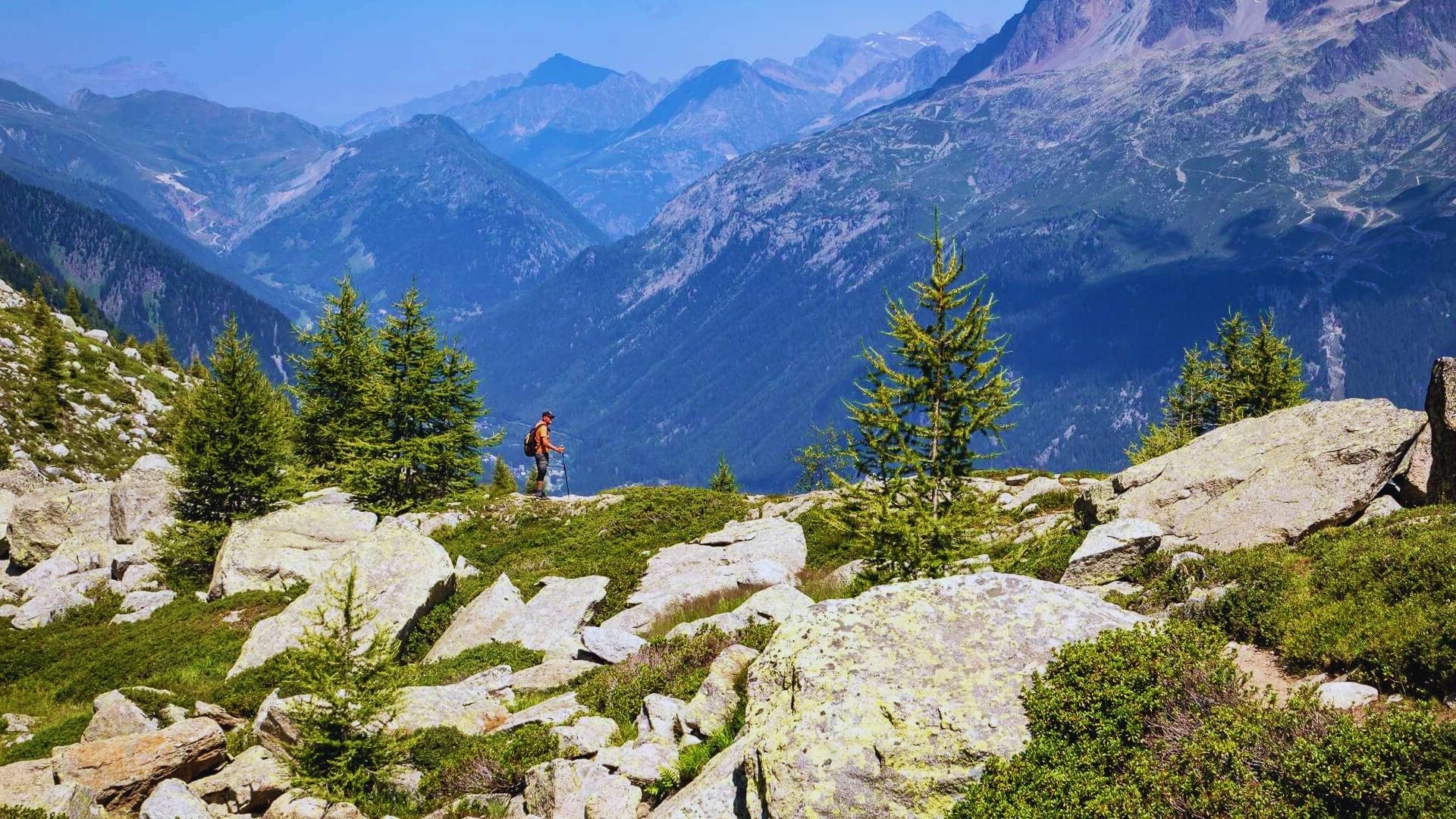 A man stands on a rocky hill, gazing at the expansive mountains in the distance.