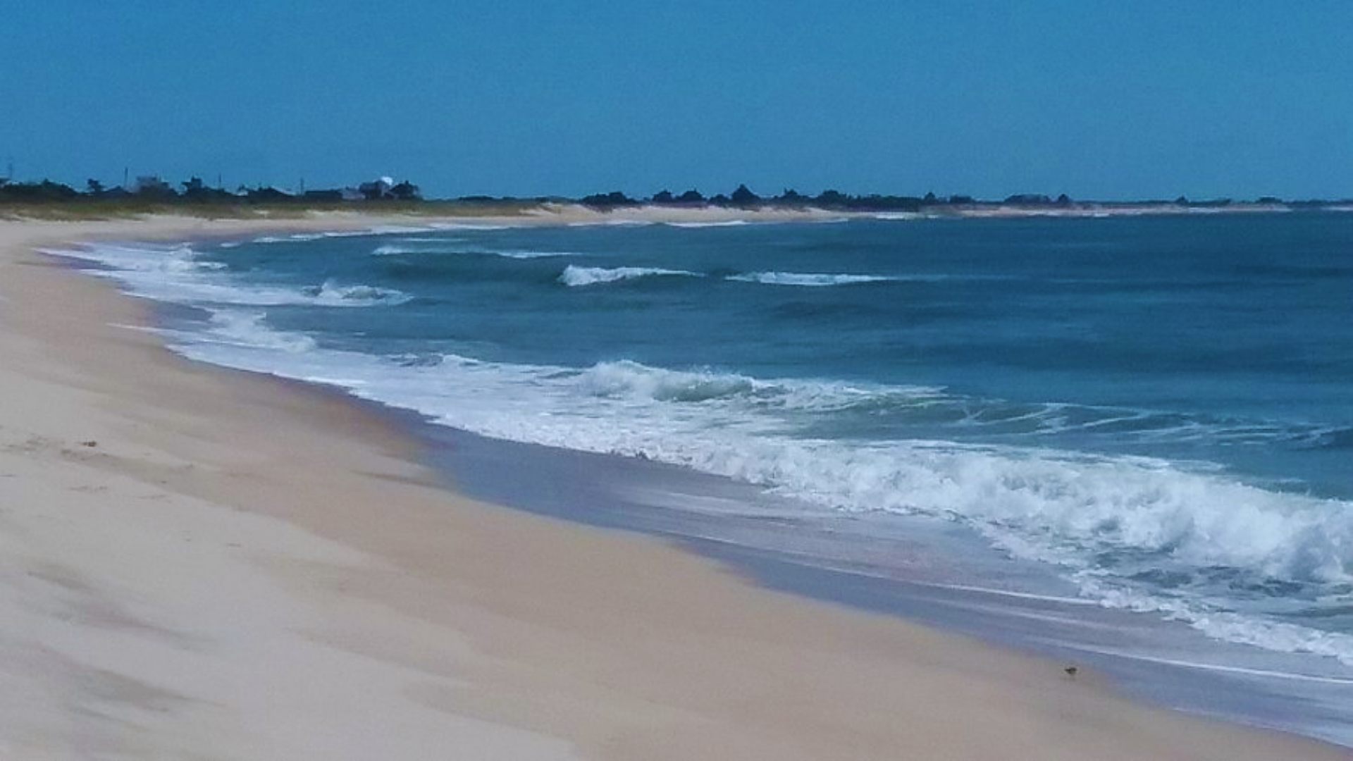 A sandy beach with gentle waves and a distant lighthouse under a clear blue sky.