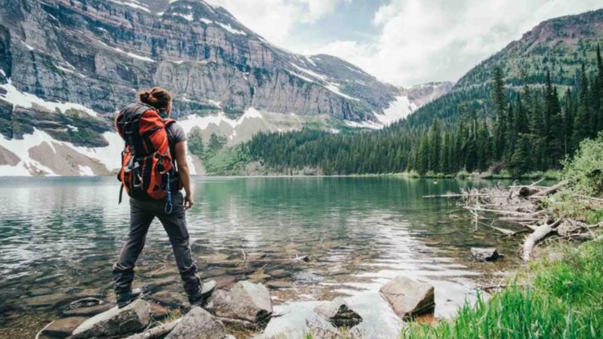A person with a backpack is perched on a rock, admiring a peaceful lake and the scenic landscape beyond.