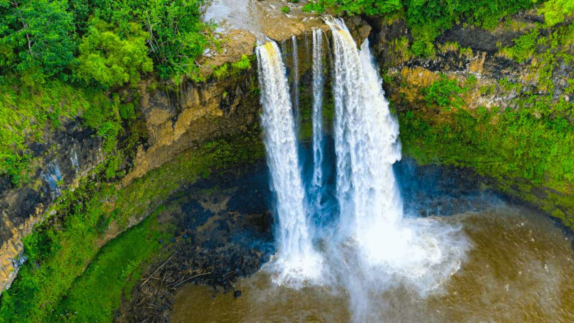 Aerial view of a waterfall cascading down a rocky cliff into a swirling pool. Lush green foliage surrounds the scene, creating a vibrant, tranquil setting.