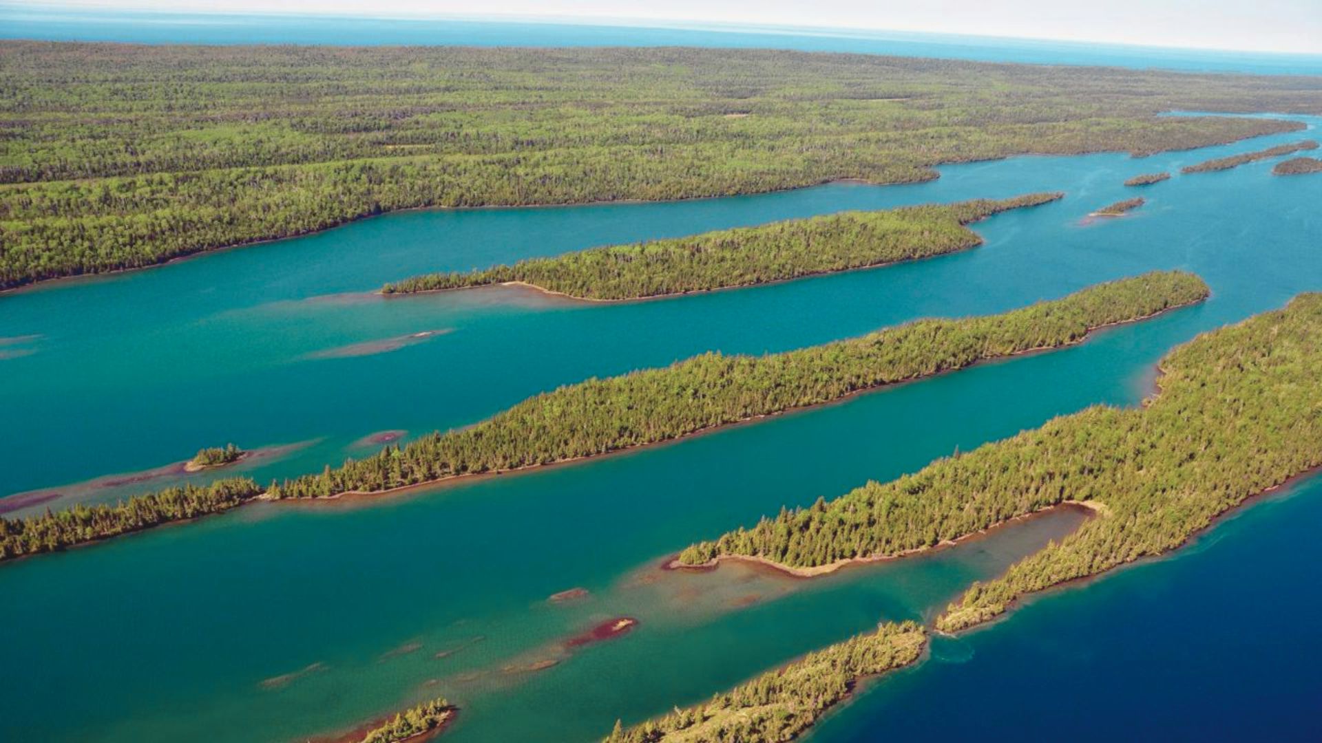 Aerial view of Isle Royale National Park, showcasing narrow, forested islands surrounded by calm, turquoise water. The landscape conveys a serene, natural beauty.