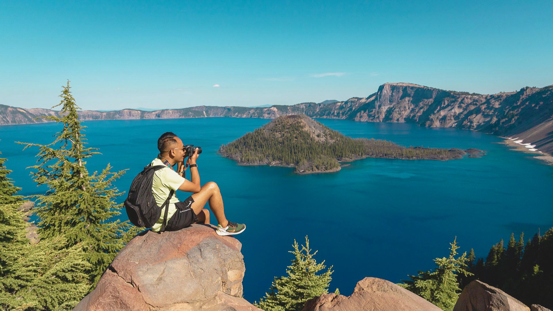 A person sits on a rock edge, capturing a photo of a blue lake and island surrounded by forested mountains. The scene conveys tranquility and adventure.