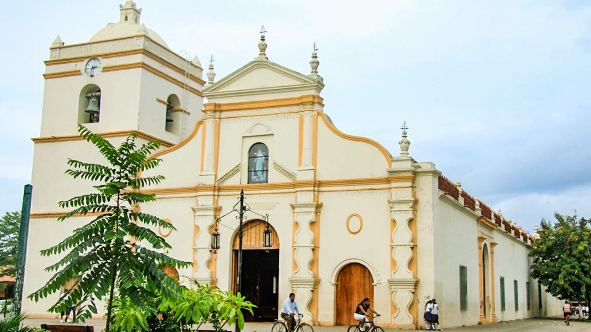 Alt text: "Historic church with a tall clock tower features ornate details and arched doorways. People pass by on bicycles, with overcast skies above."
