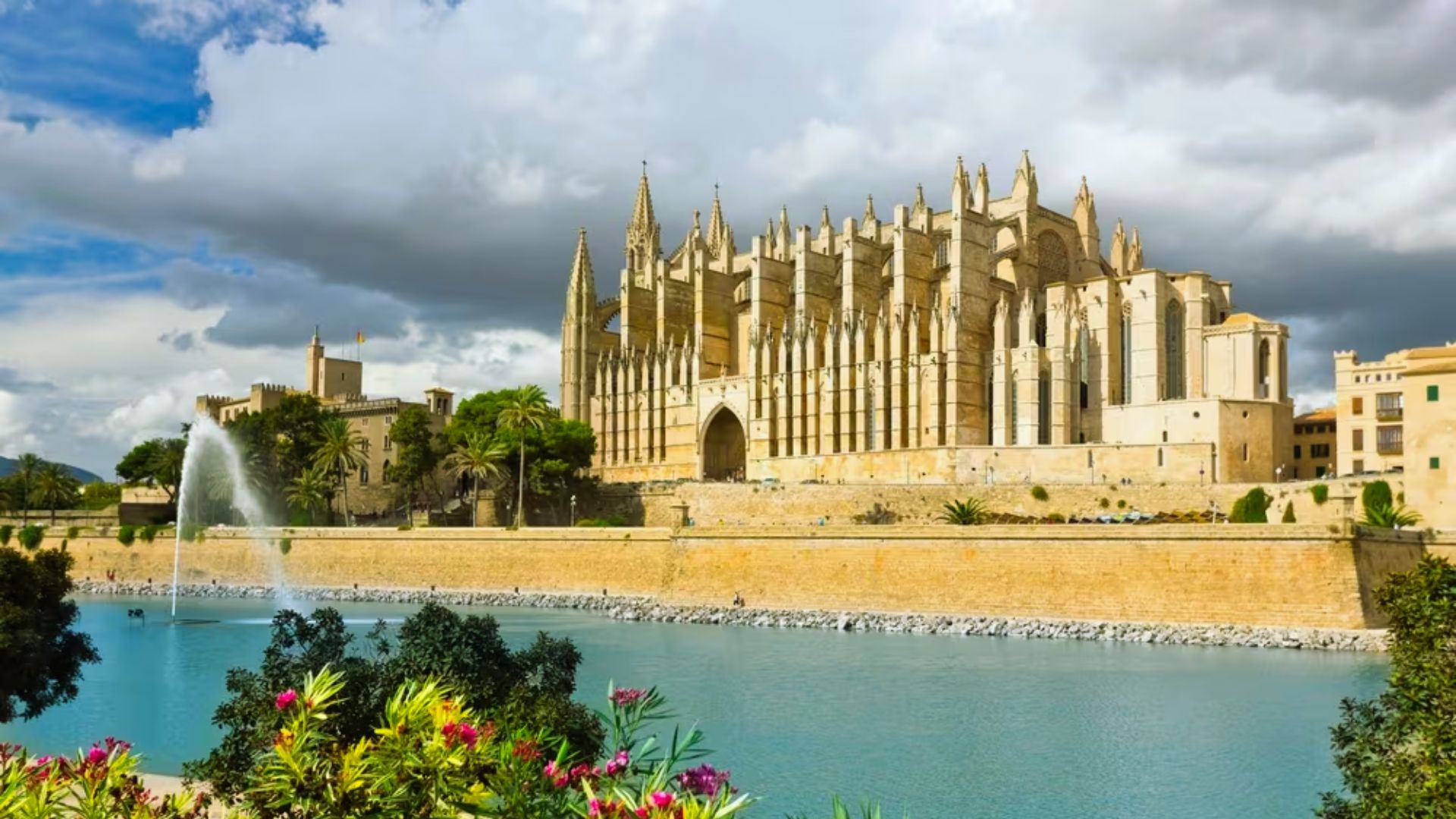 Historic cathedral with ornate spires overlooks a turquoise pool with a central fountain, framed by lush greenery under dramatic clouds.
