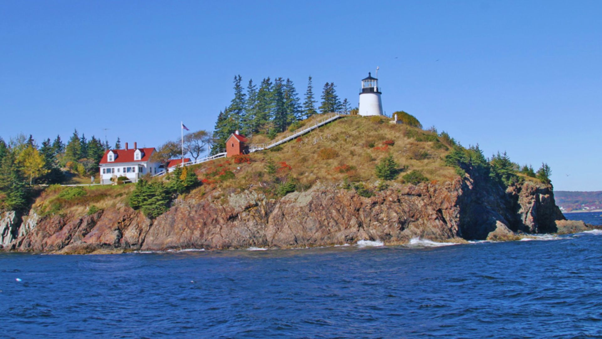 Remote island with a white lighthouse atop rocky cliffs, surrounded by lush greenery and trees. A cozy house sits nearby. The sea is calm under a clear blue sky.