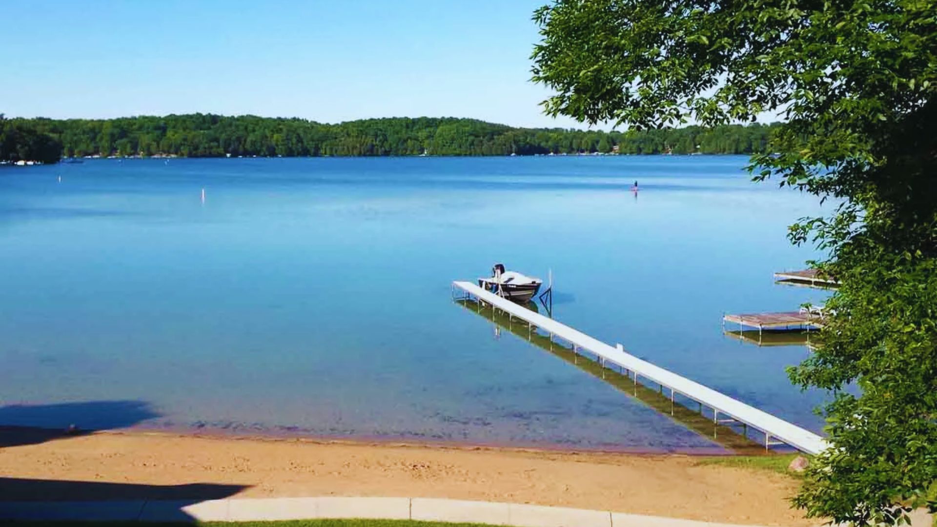 A serene lakeside scene with a sandy beach, a long dock extending into calm blue water, and green trees in the background. A single boat is moored at the dock.