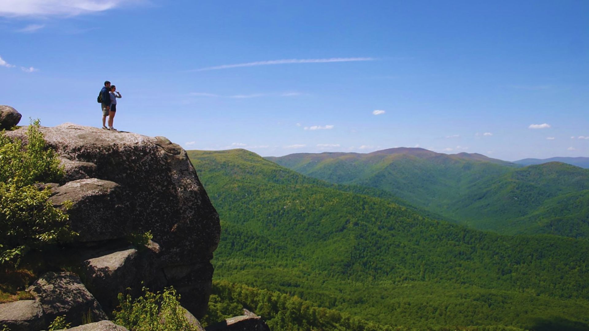 A lone hiker stands on a large rocky cliff, overlooking vast, lush green mountains under a clear blue sky, capturing a sense of adventure and freedom.