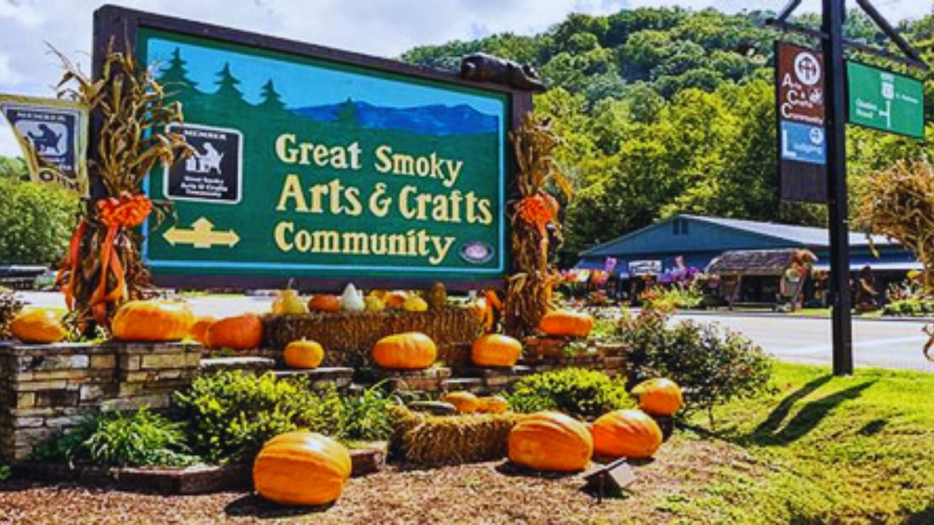 Sign for "Great Smoky Arts & Crafts Community" adorned with pumpkins, hay bales, and cornstalks. Lush green forested hills in the background.
