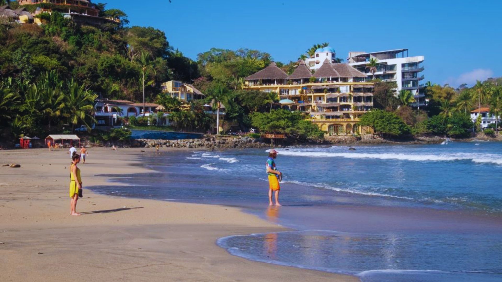 Two people in bright swimwear stand on a sandy beach under a clear blue sky, with gentle waves nearby and lush, green hills and buildings in the background.