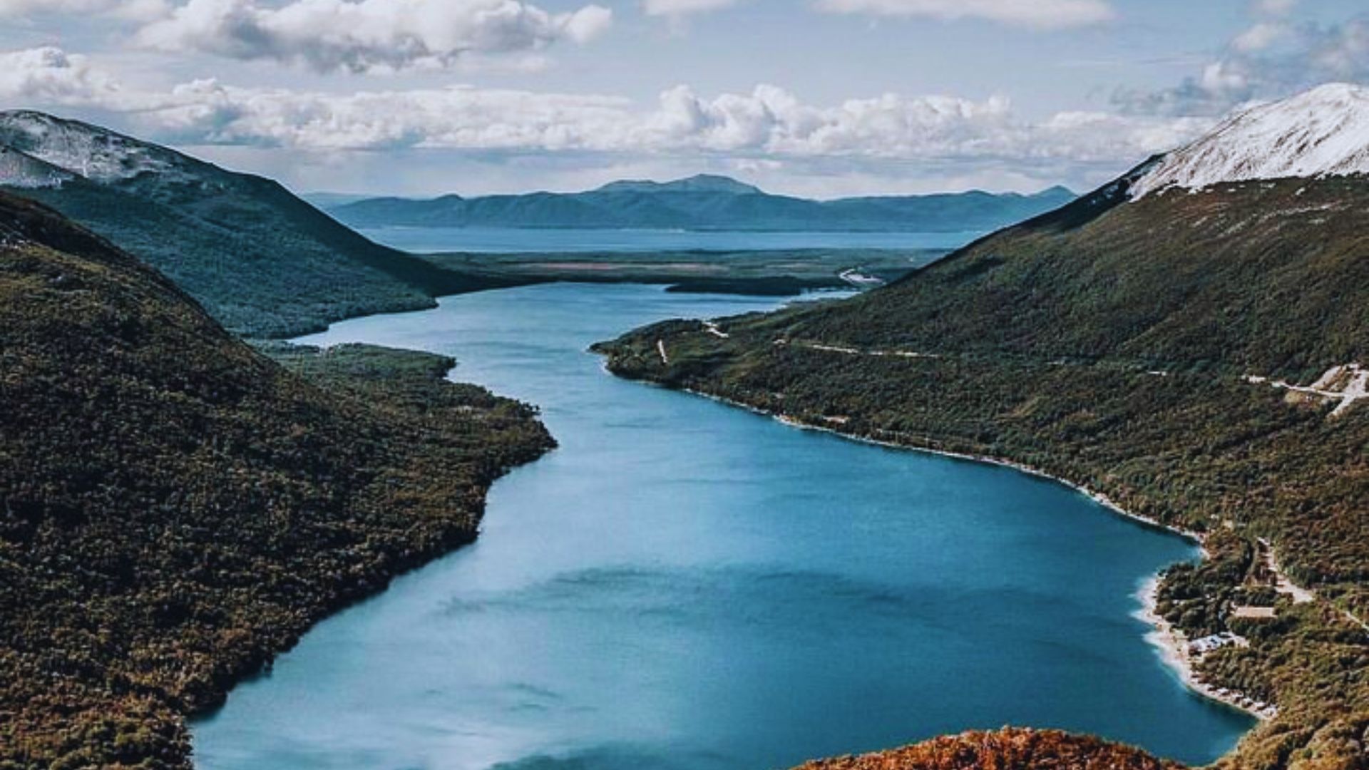 A serene, wide river curves between lush, green mountains under a partly cloudy sky. Snow dusts the peaks, conveying a sense of peace and grandeur.