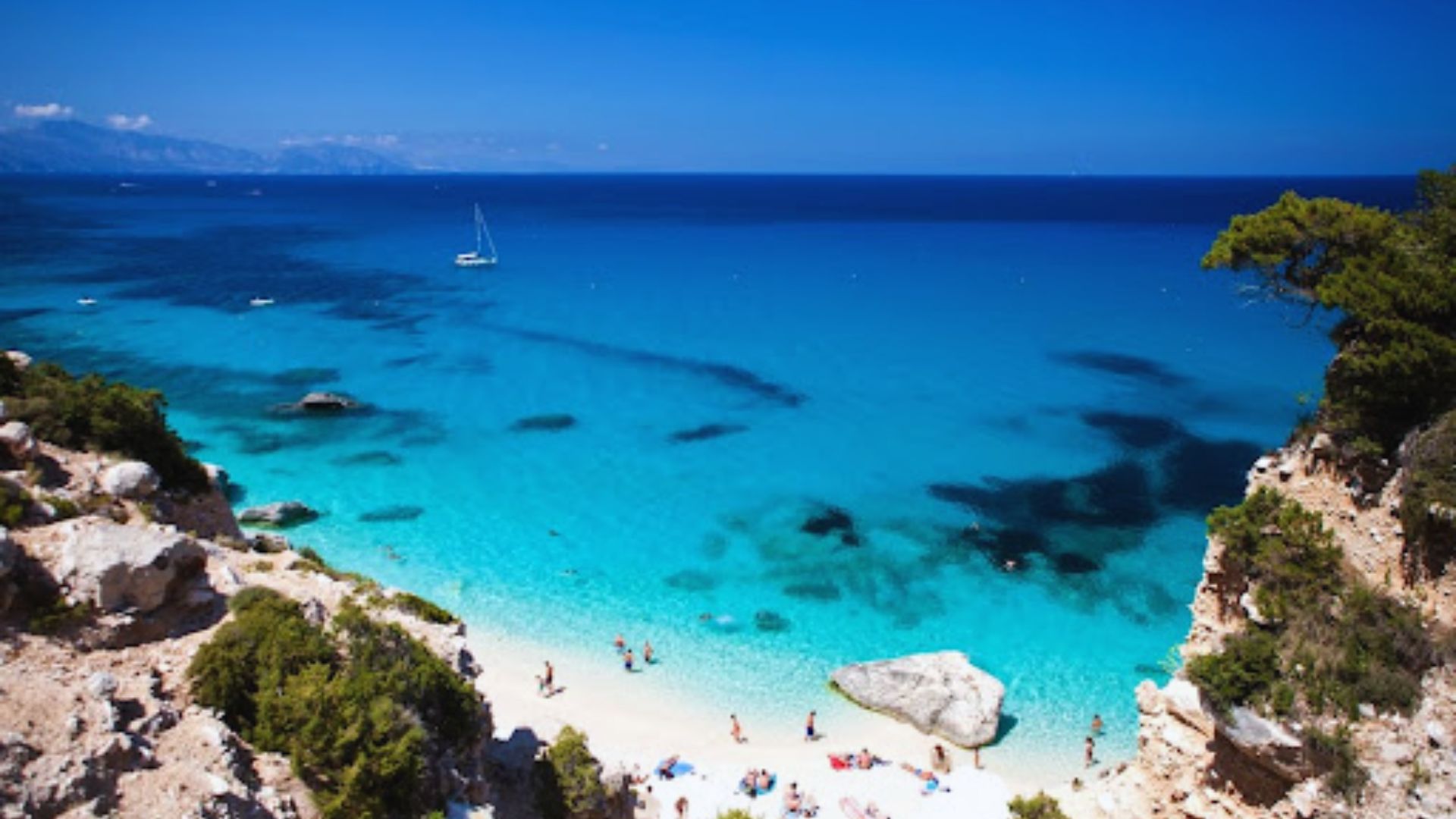 A breathtaking view of a vibrant turquoise beach framed by rocky cliffs and lush greenery. A few people enjoy the sun, and a sailboat floats nearby.