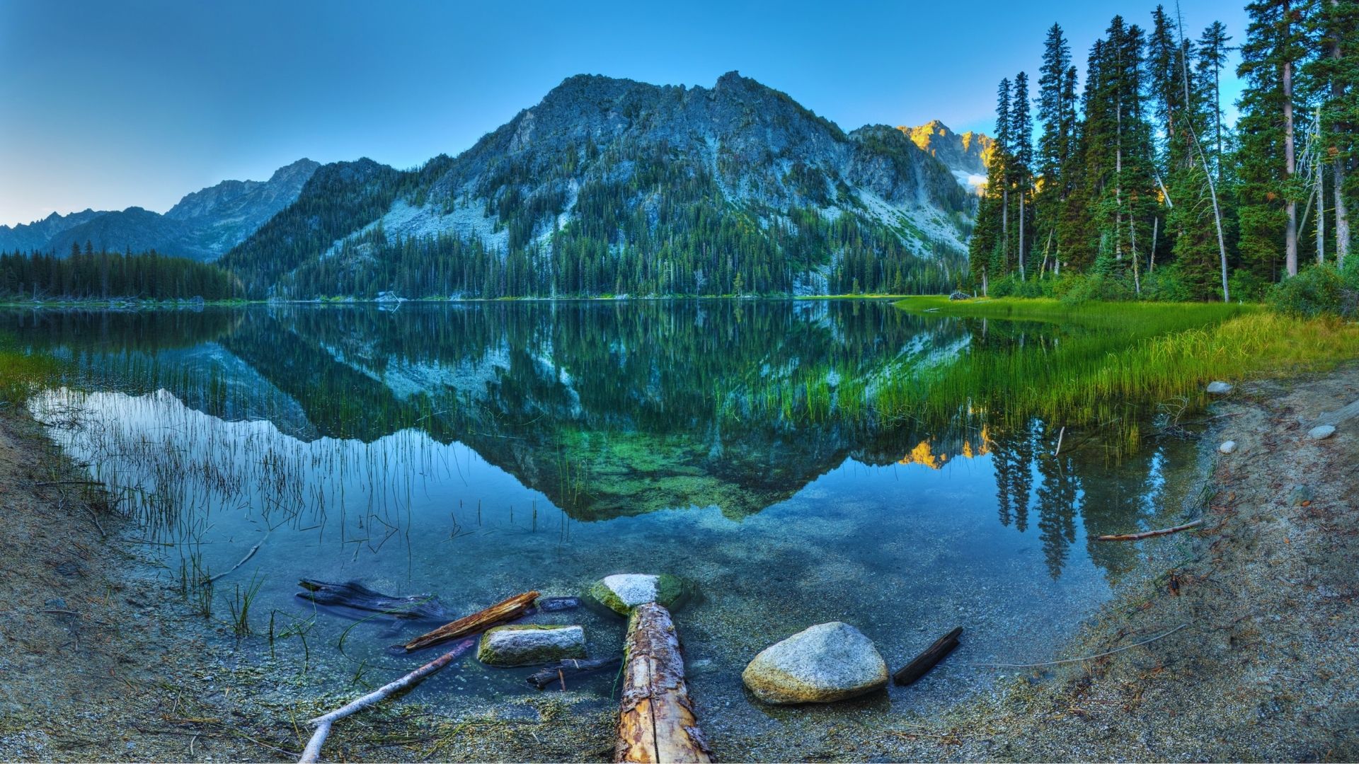 A serene mountain lake bordered by lush trees and rugged rocks under a clear blue sky.