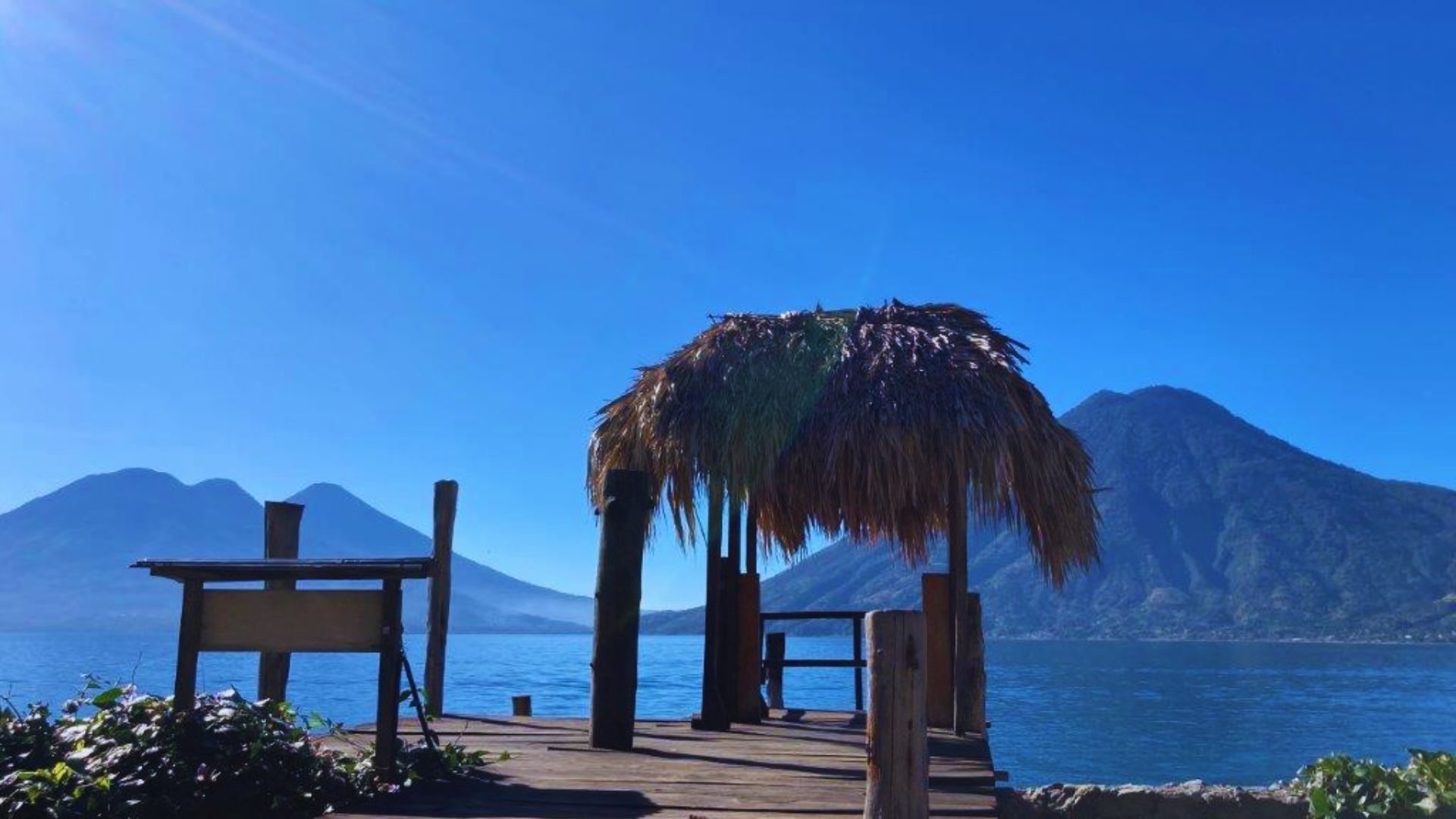 A wooden dock with a thatched roof extends over water, framed by mountains in the background under a clear sky.