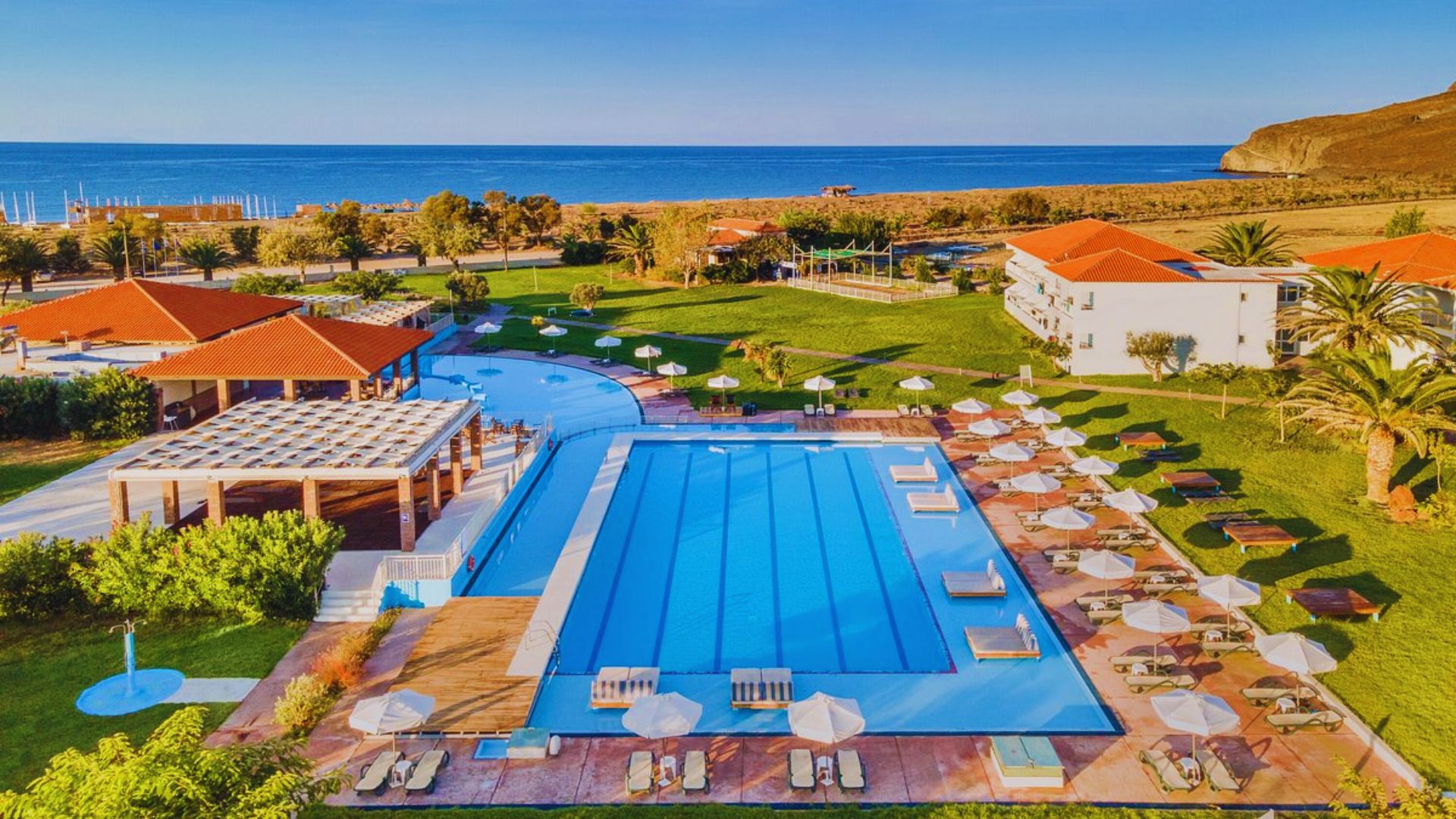 Aerial view of a luxury resort featuring a large, rectangular swimming pool surrounded by lounge chairs and parasols. The resort overlooks a serene beach with calm, blue ocean waves. Red-roofed buildings and lush greenery add to the tranquil atmosphere.